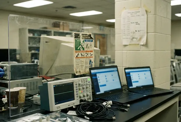 Laboratory equipment close-up, oscilloscope display showing idle waveform beside a cluttered workstation with tangled USB